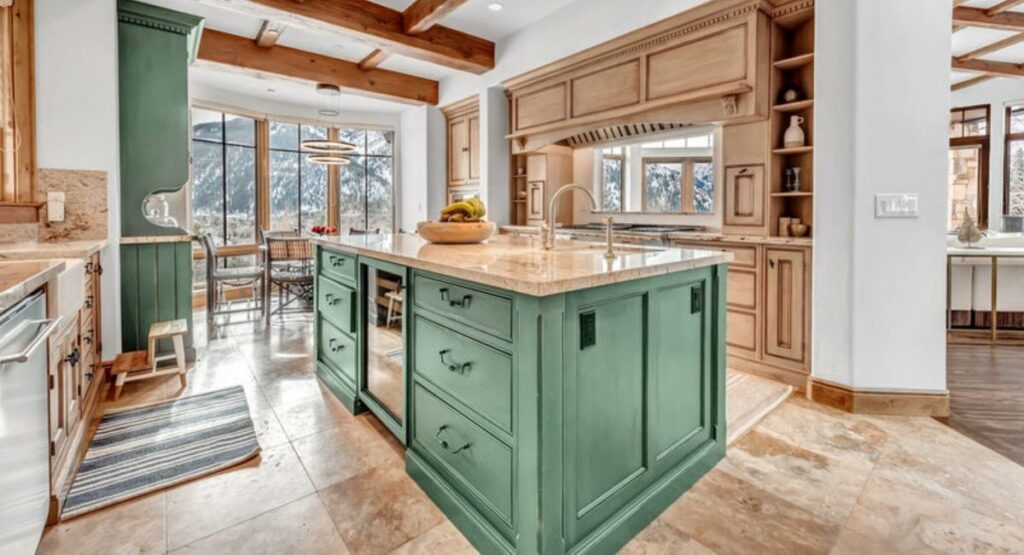 A spacious kitchen with a rustic vibe features a central green island with a fruit bowl, wooden beams, beige cabinets, and large windows showing snowy mountains.